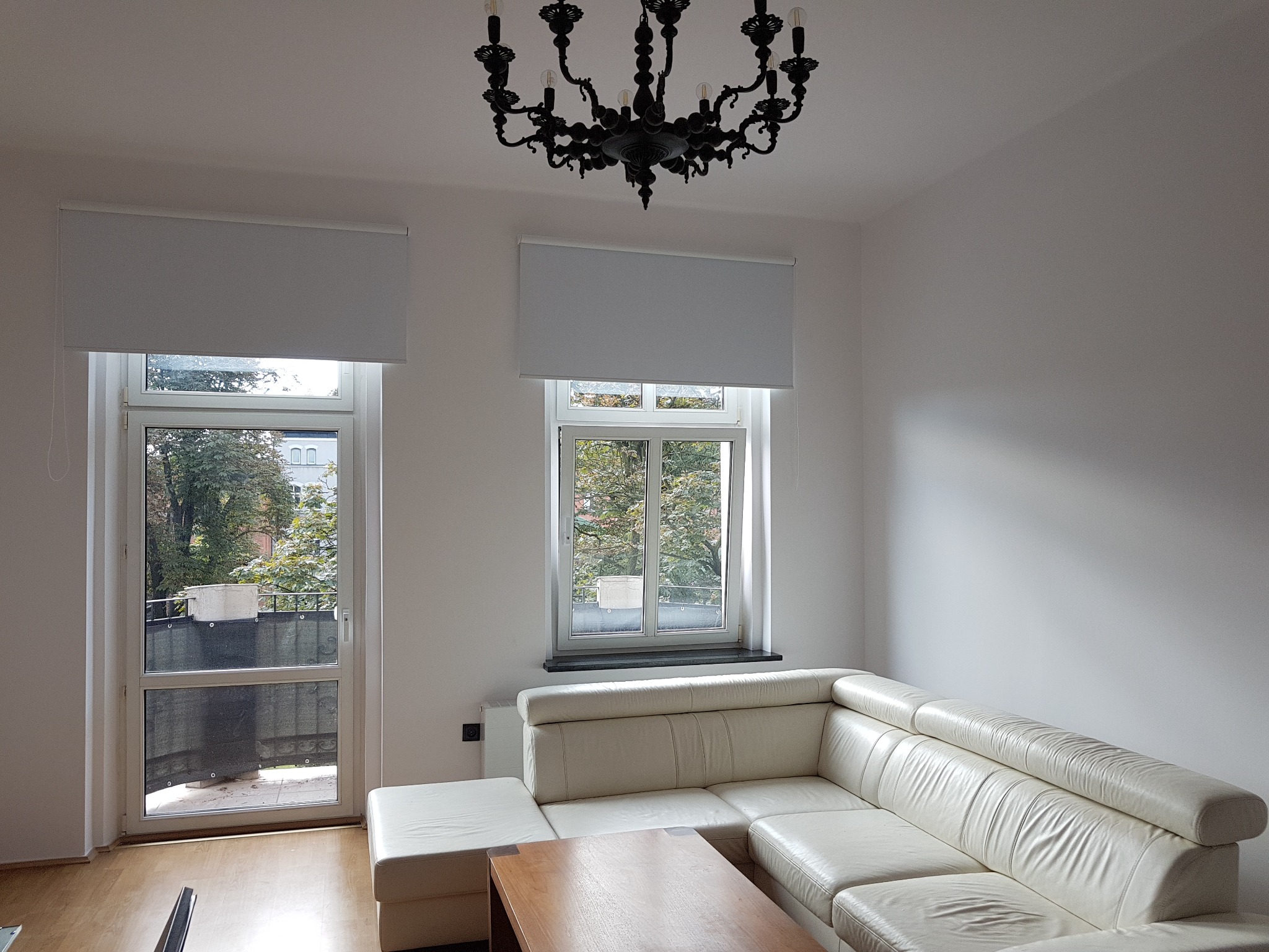 Jasne wnętrze pokoju z białym narożnikiem, drewnianym stolikiem kawowym i oknami z roletami, oświetlone czarnym, ozdobnym żyrandolem.