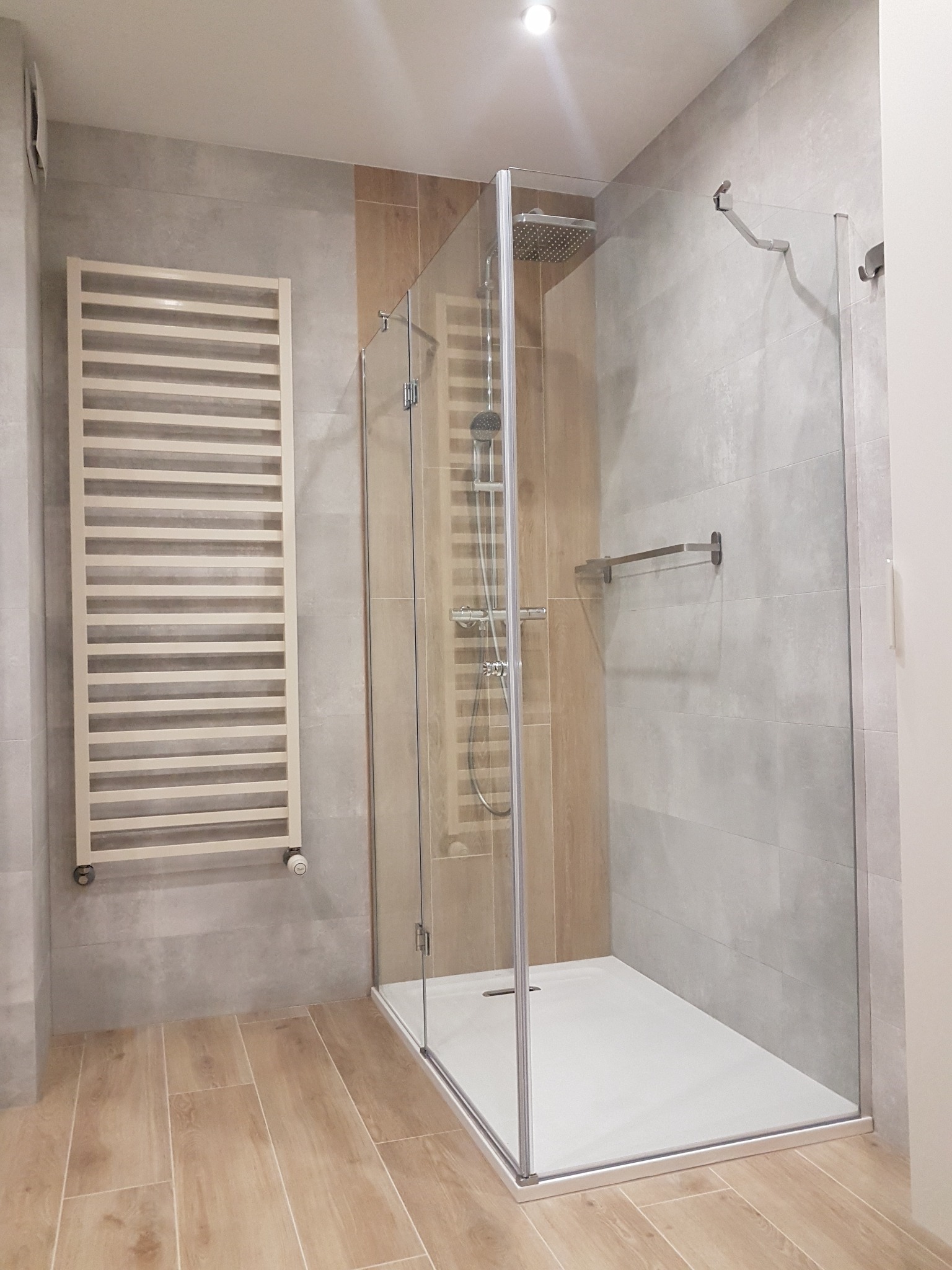 Nowoczesna kabina prysznicowa z deszczownicą, szklanymi ściankami i białym brodzikiem, obok grzejnik drabinkowy na szarej ścianie z imitacją betonu, podłoga wyłożona panelami imitującymi drewno.