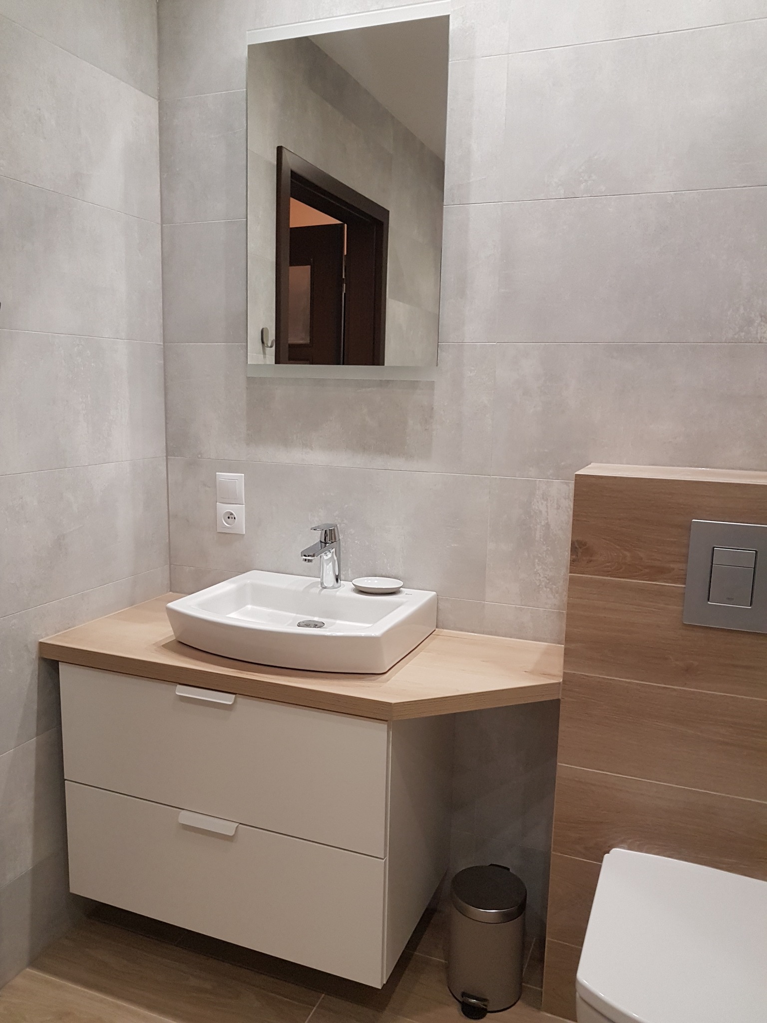 Nowoczesna łazienka z umywalką nablatową, drewnianym blatem, szarą armaturą i płytkami imitującymi beton, widok na lustro i fragment toalety.