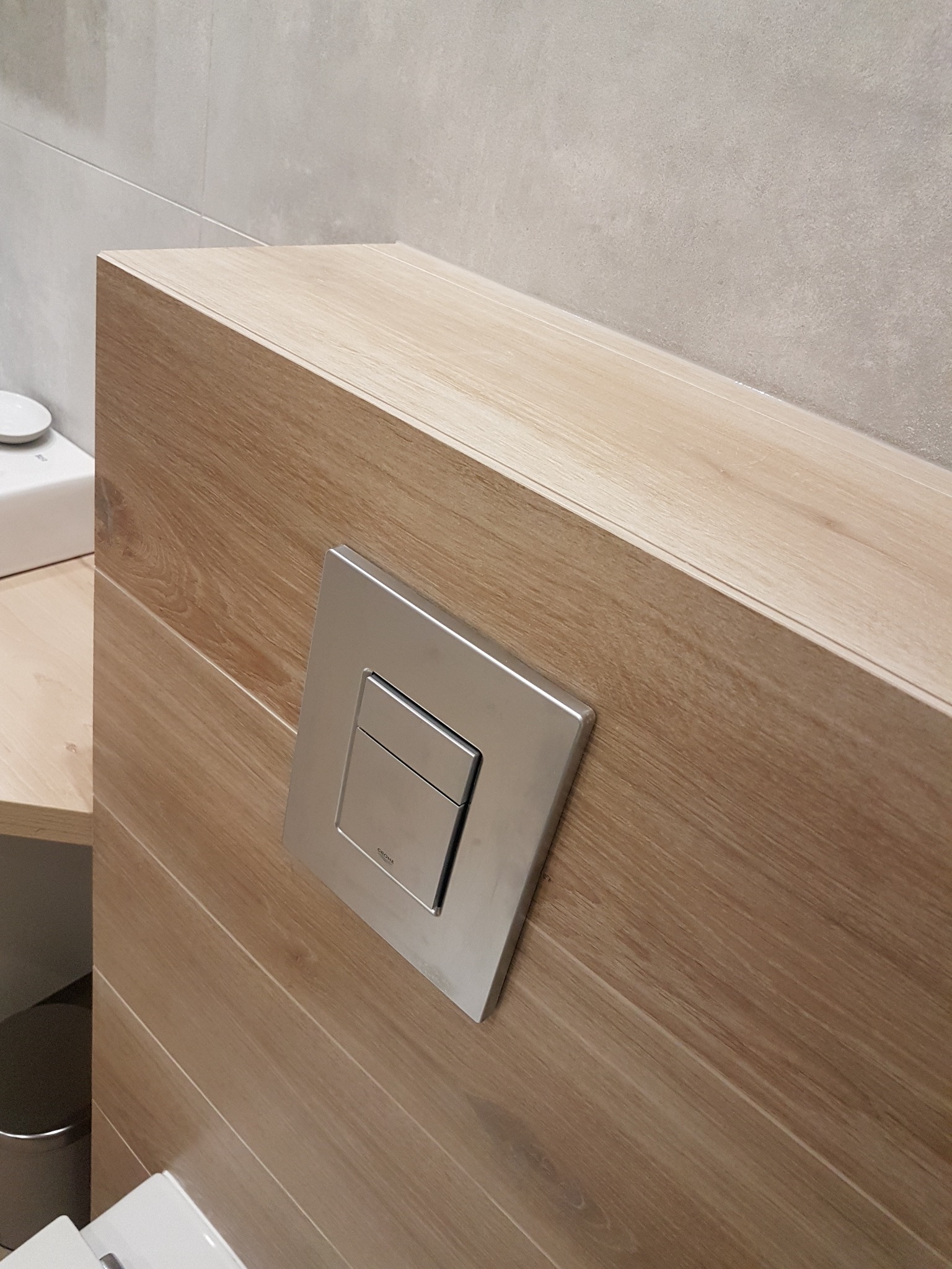 Szczegół zabudowy stelaża WC z drewnopodobną okładziną i chromowanym przyciskiem spłukującym marki Grohe, na tle jasnoszarych płytek ceramicznych.