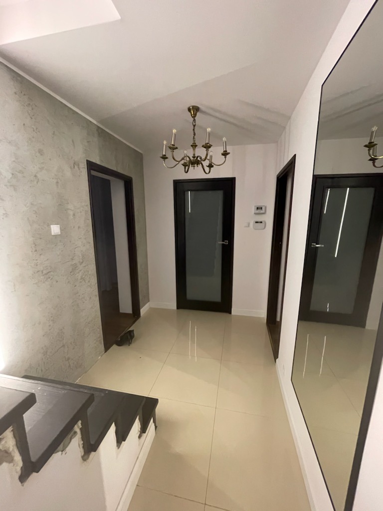 Wykończony korytarz z beżowymi płytkami podłogowymi, szarą ścianą z efektem betonu, ciemnymi drzwiami z szybami, lustrem i metalowym żyrandolem.