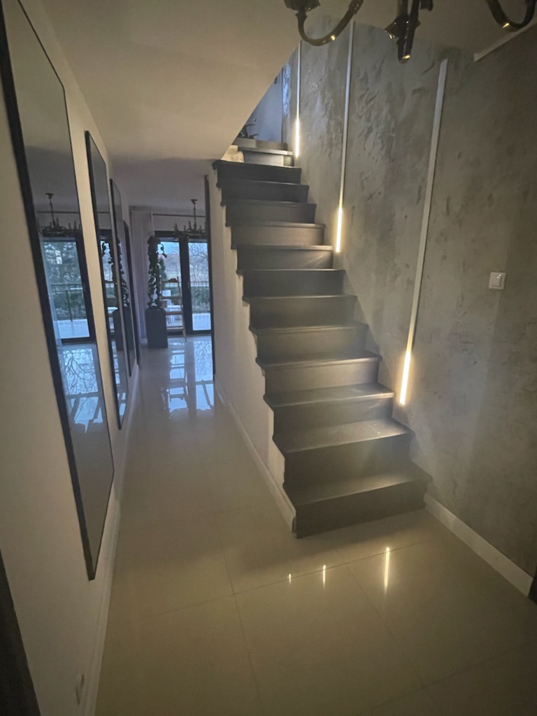 Nowoczesne schody z podświetleniem LED wzdłuż ściany o fakturze betonu, widok z dołu na korytarz z lustrami i przeszklonymi drzwiami.