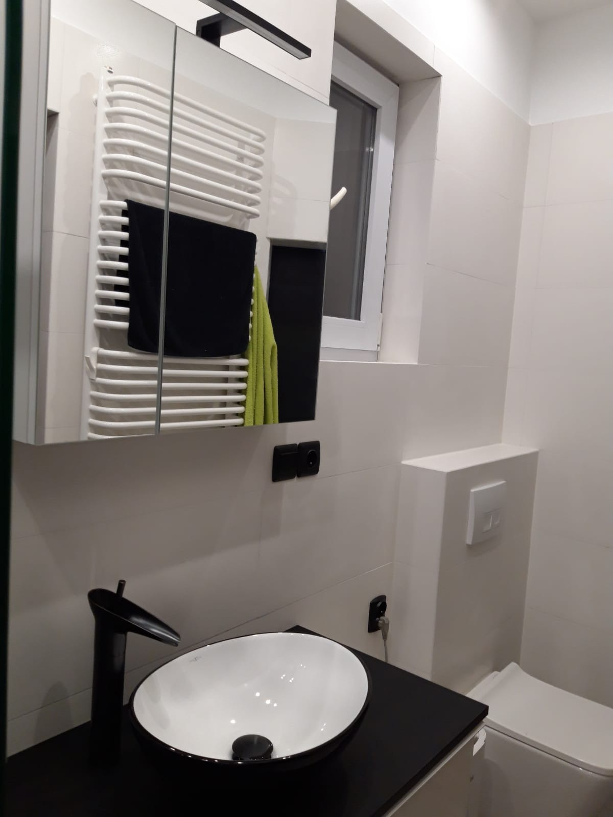 Nowoczesna łazienka z białą umywalką nablatową, czarną armaturą i grzejnikiem drabinkowym z czarnym i zielonym ręcznikiem, widoczne gniazdka elektryczne i fragment toalety.
