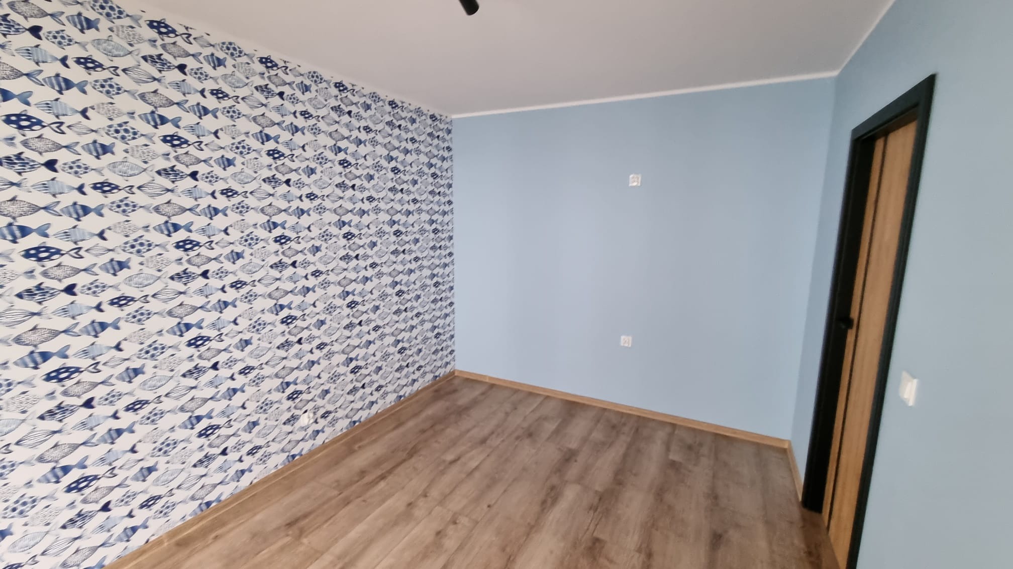 Pokój po remoncie: ściana z tapetą w niebieskie rybki, ściana pomalowana na jasnoniebiesko, panele podłogowe w odcieniach brązu, ciemna futryna drzwiowa.