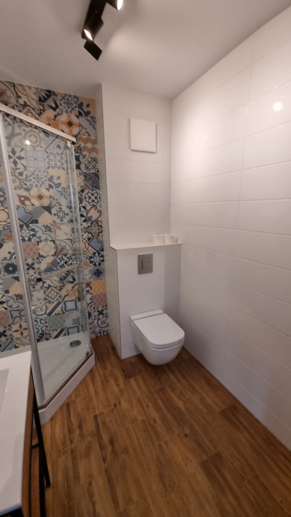 Wykończona łazienka z drewnopodobną podłogą, białą armaturą, kabiną prysznicową z wzorzystymi płytkami oraz minimalistyczną, podtynkową spłuczką.