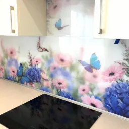 Szklany panel kuchenny z motywem kwiatów i motyli, zamontowany nad płytą indukcyjną, widoczne kremowe szafki.