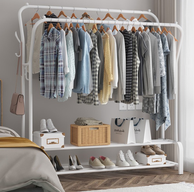 Dwupoziomowy, biały metalowy wieszak na ubrania z drewnianymi wieszakami, różnorodną odzieżą, pudełkami, torbami i obuwiem na półkach, ustawiony w sypialni obok łóżka.