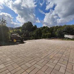 Nowo ułożona, beżowo-brązowa kostka brukowa w jodełkę na podjeździe, z widocznym fragmentem trawnika, przyczepką i stosem płyt chodnikowych w tle pod błękitnym niebem z białymi chmurami.