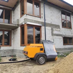 Maszyna do tynkowania na tle surowego budynku z oknami w drewnianych ramach, widoczne ślady zaprawy między bloczkami.