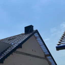 Budowa domu w trakcie: widok na dach z czarną dachówką i kominem, ściany z białego pustaka, niebieska membrana dachowa, drewniana konstrukcja dachu sąsiedniego budynku, błękitne niebo i smuga...