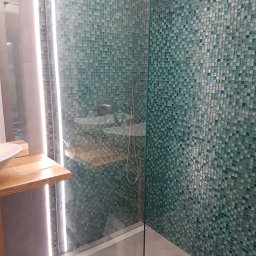 PROELEBUD - Szklana kabina prysznicowa z mozaiką w odcieniach zieleni i błękitu, oświetlenie LED wzdłuż krawędzi kabiny, drewniany blat umywalki z białą umywalką nablatową, szara podłoga i ściany.