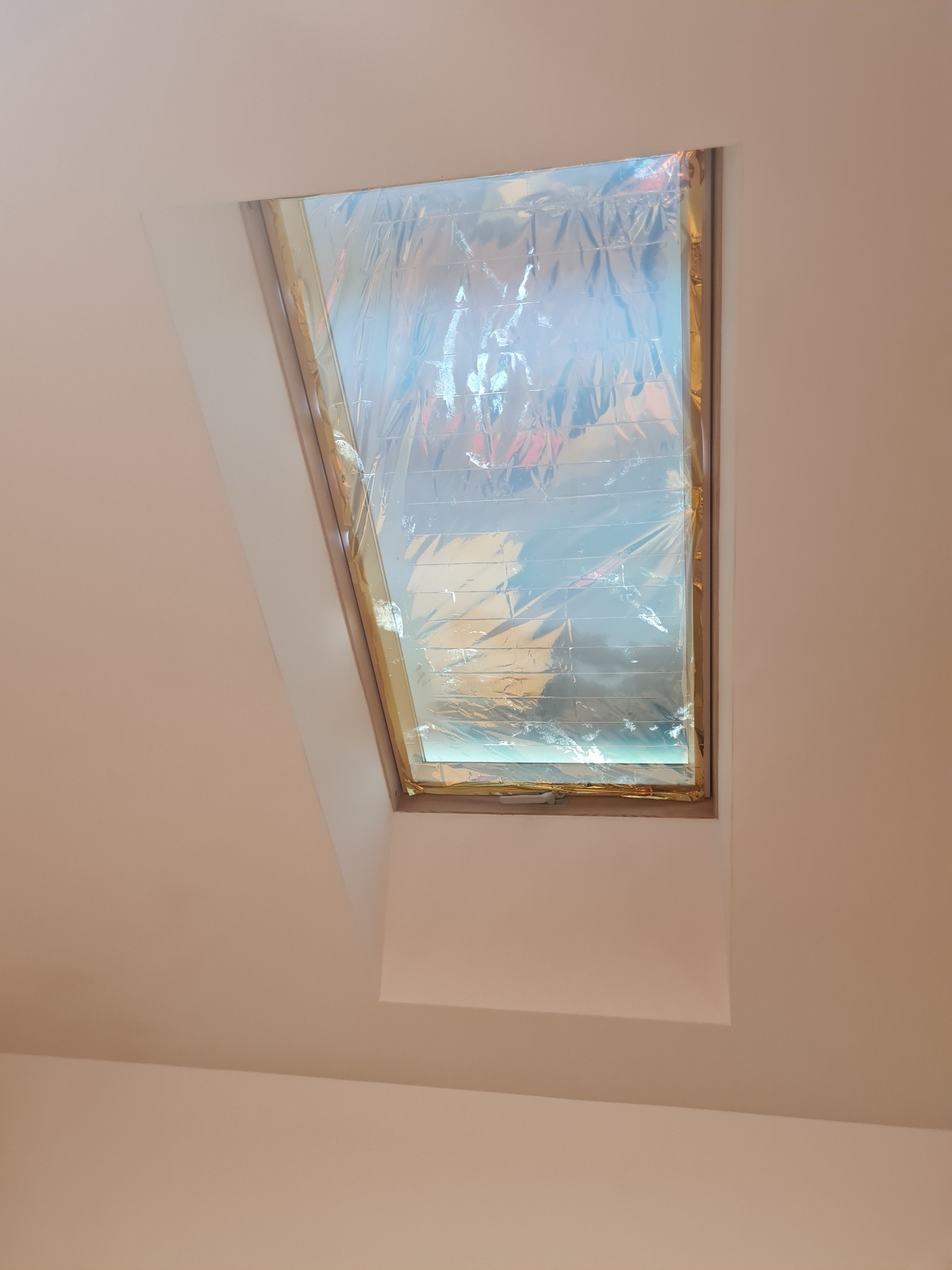 Okno dachowe zabezpieczone folią ochronną podczas prac remontowych, widok od wewnątrz, jasne ściany.