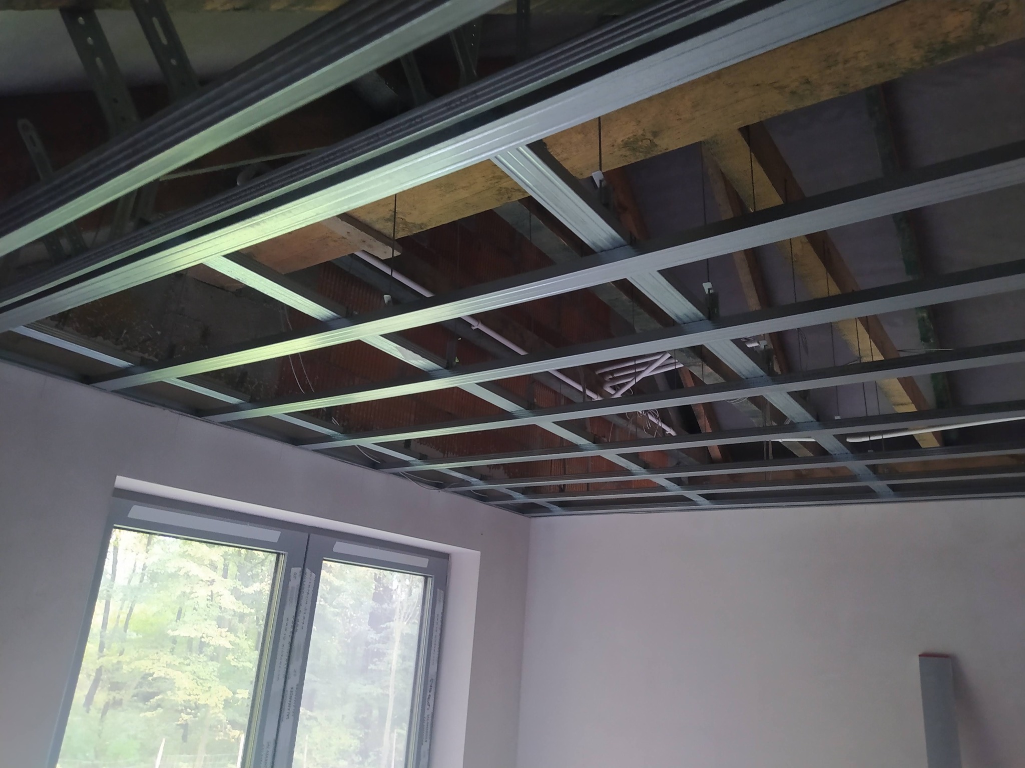 Stelaż sufitu podwieszanego z widocznymi elementami konstrukcyjnymi i przewodami, okno z widokiem na zieleń w tle, ściany w kolorze białym.