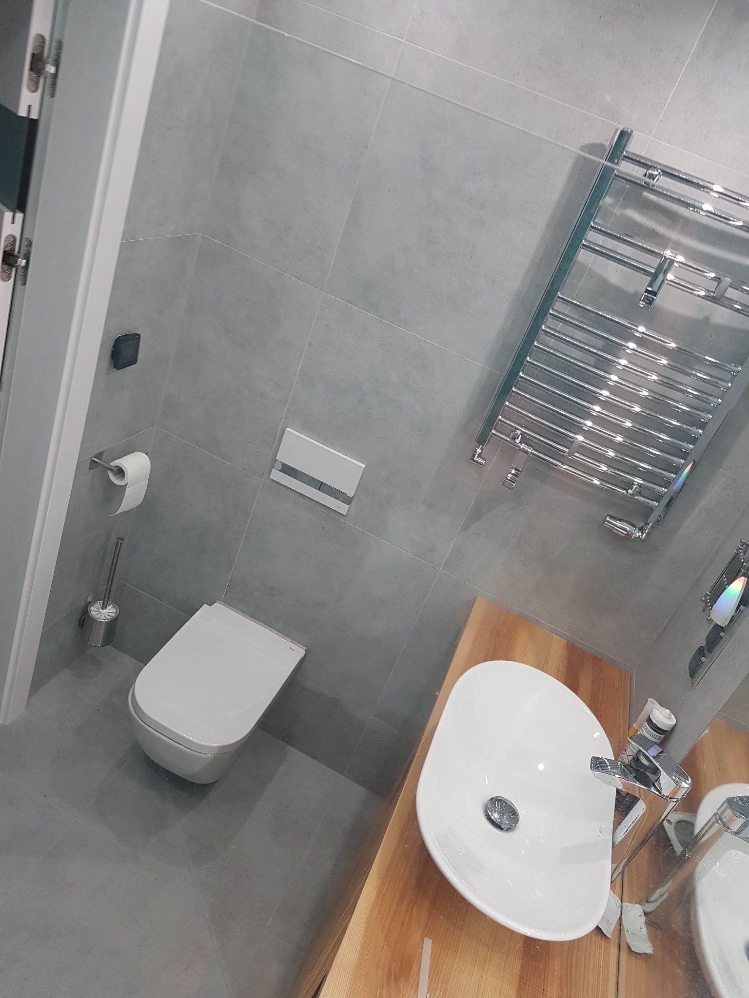 Nowoczesna łazienka z szarymi płytkami, wiszącą toaletą, drewnianym blatem pod umywalką i chromowanym grzejnikiem drabinkowym.