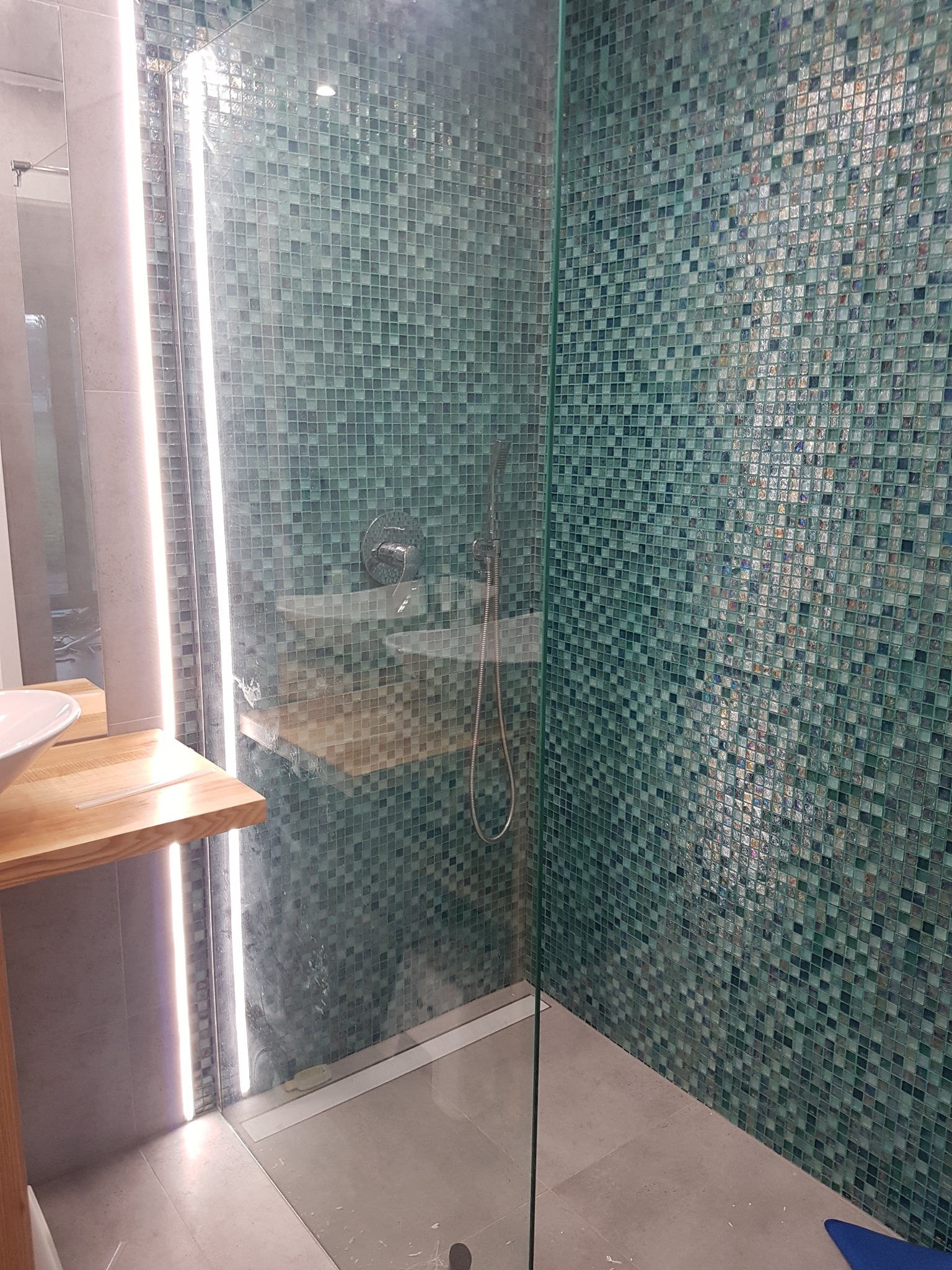 Szklana kabina prysznicowa z mozaiką w odcieniach zieleni i błękitu, oświetlenie LED wzdłuż krawędzi kabiny, drewniany blat umywalki z białą umywalką nablatową, szara podłoga i ściany.