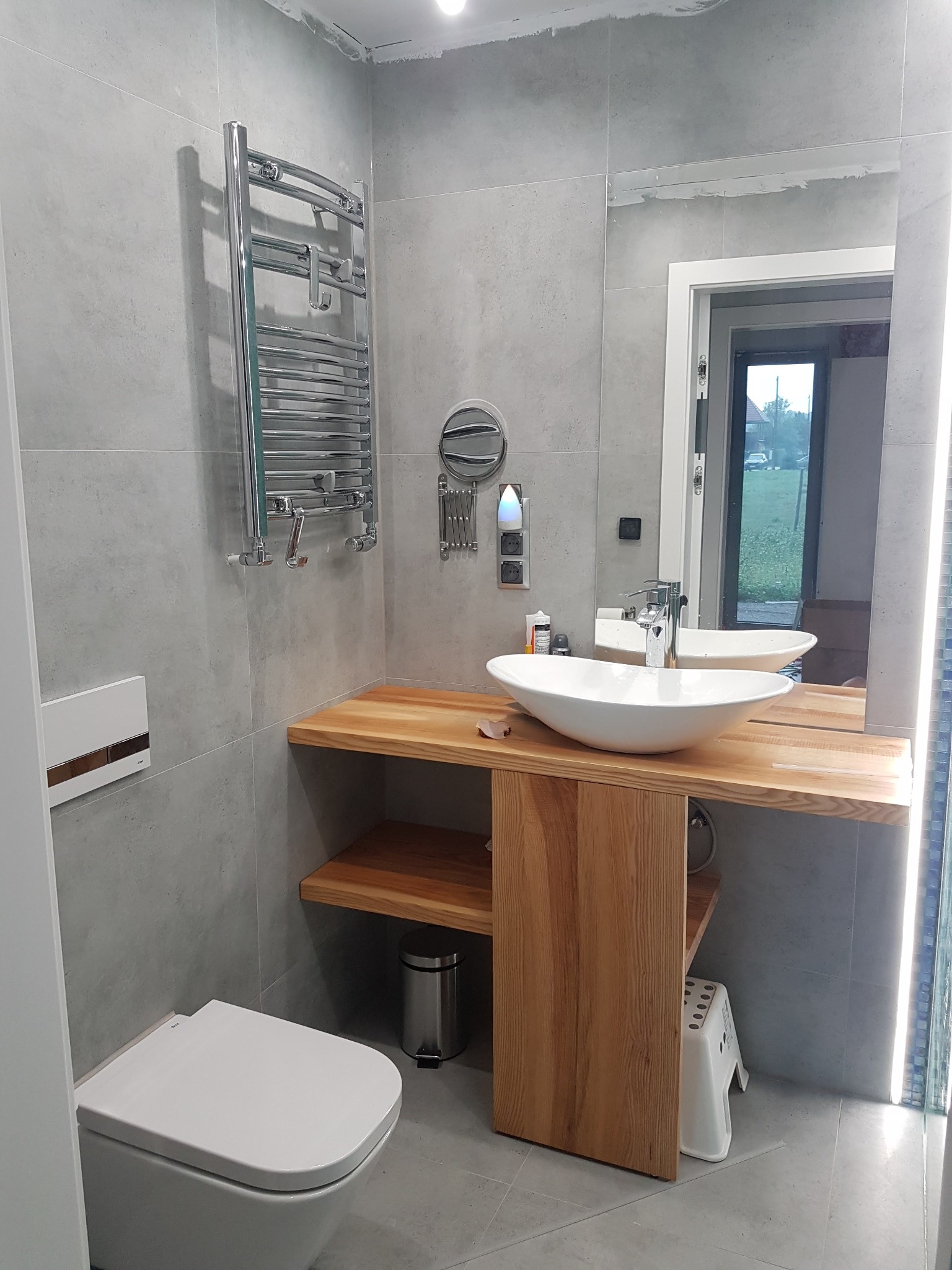 Nowoczesna łazienka w szarościach z drewnianym blatem pod umywalką, wiszącym grzejnikiem drabinkowym i podświetlanym lustrem. Widoczna toaleta i mały stołek pod blatem.