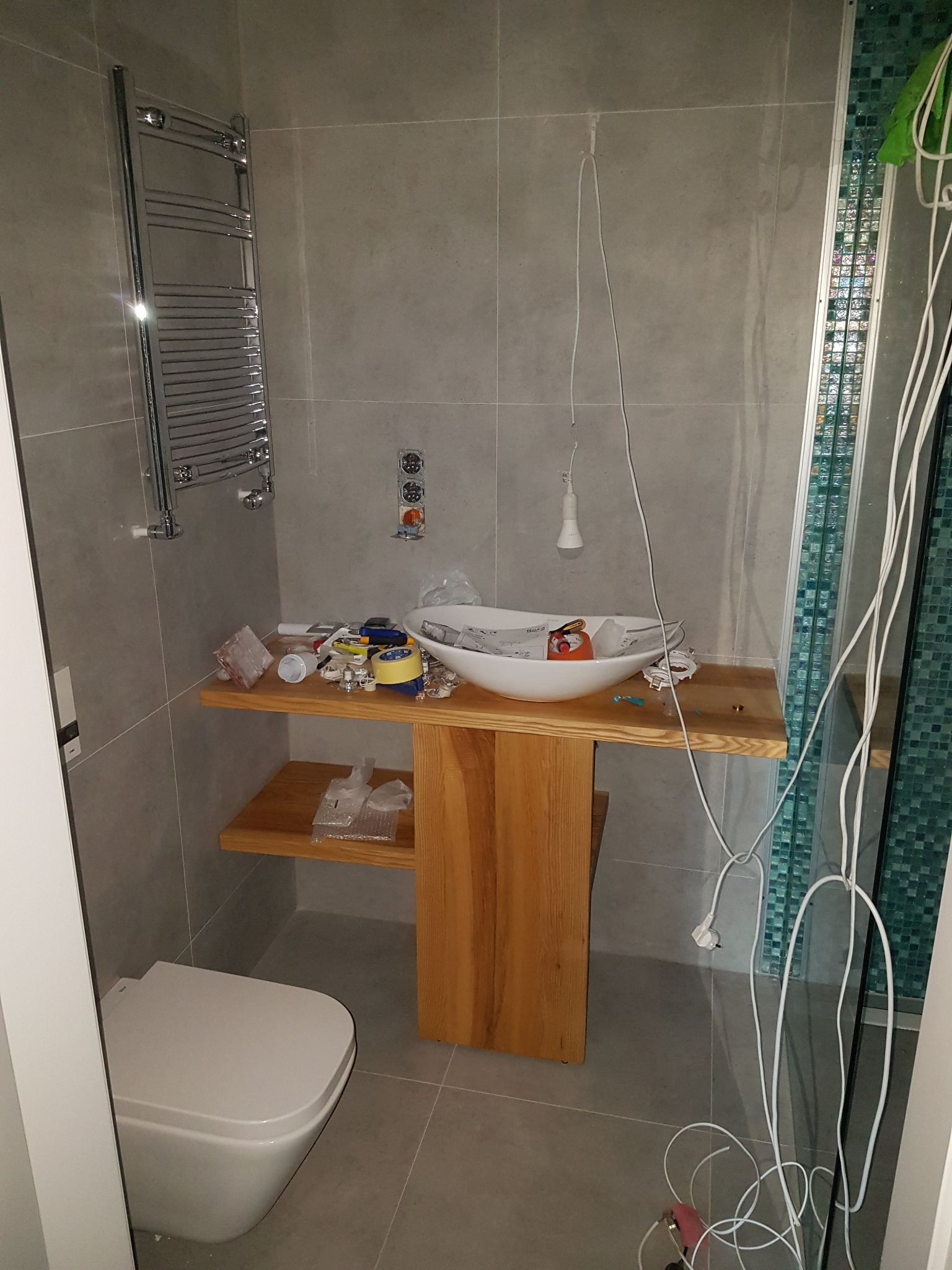 Łazienka w trakcie prac wykończeniowych: umywalka nablatowa na drewnianym blacie z półką, grzejnik drabinkowy na ścianie, wisząca żarówka na kablu, narzędzia i materiały budowlane na blacie...