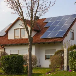 Dom jednorodzinny z czerwonym dachem i panelami słonecznymi na dwóch połaciach dachu, widok z ogrodu w słoneczny dzień.