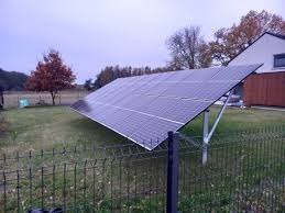 Panele fotowoltaiczne na metalowej konstrukcji ustawione na trawniku za niskim, czarnym ogrodzeniem z siatki, w tle dom jednorodzinny i drzewa pod pochmurnym niebem.