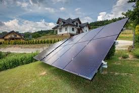 Panel słoneczny ustawiony pod kątem na zielonym trawniku przed domem jednorodzinnym z ciemnym dachem, widoczne niebo z chmurami.