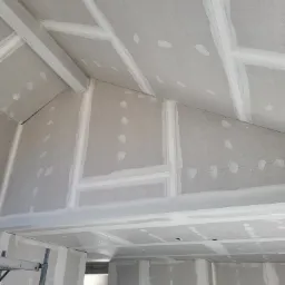 Skośny sufit i ściany poddasza w trakcie montażu płyt gipsowo-kartonowych, widoczne białe profile i łączenia płyt wypełnione szpachlą, drabina w tle.
