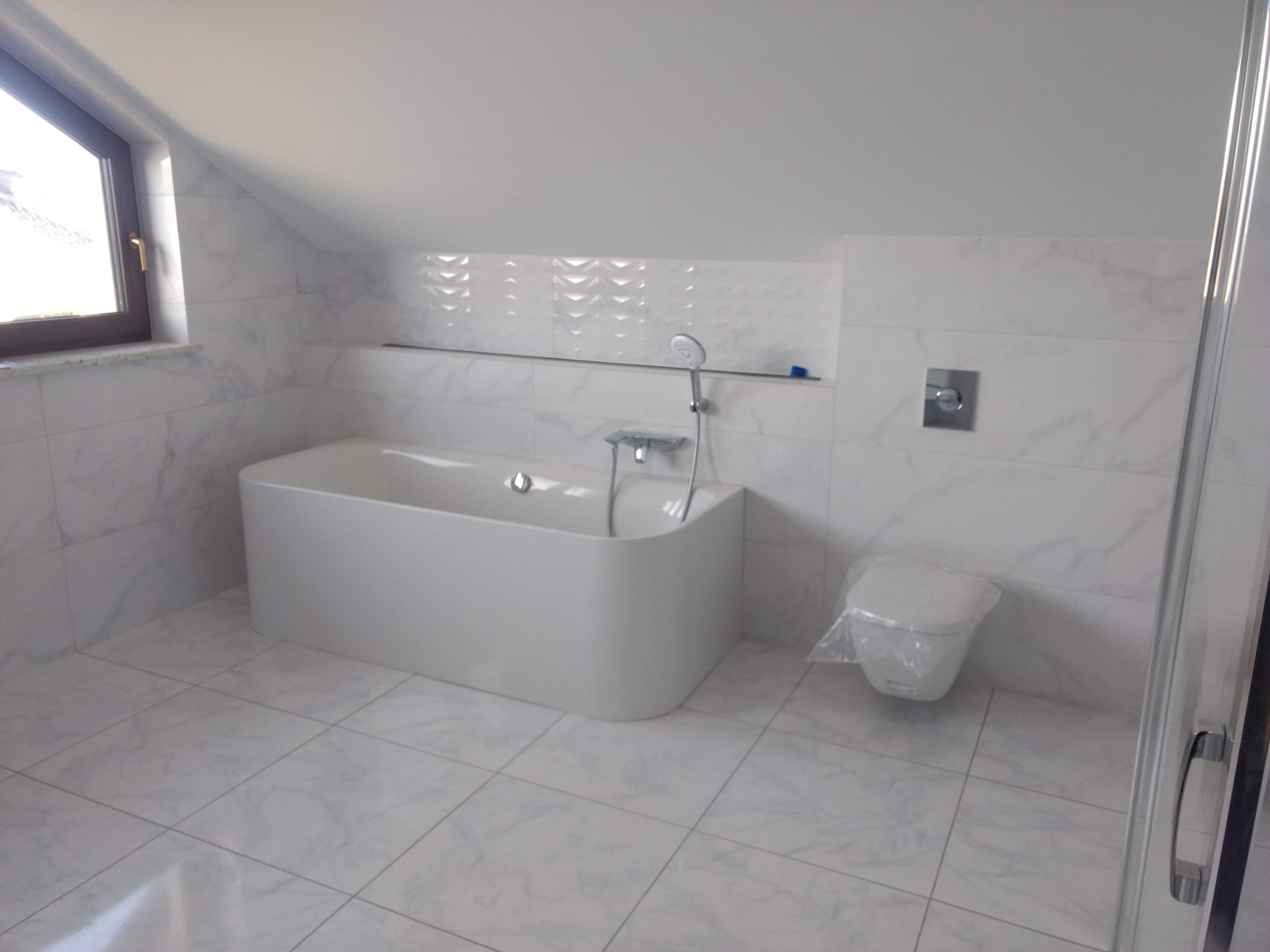 Nowoczesna łazienka na poddaszu z białą, wolnostojącą wanną, marmurowymi płytkami i toaletą w folii ochronnej przed montażem.