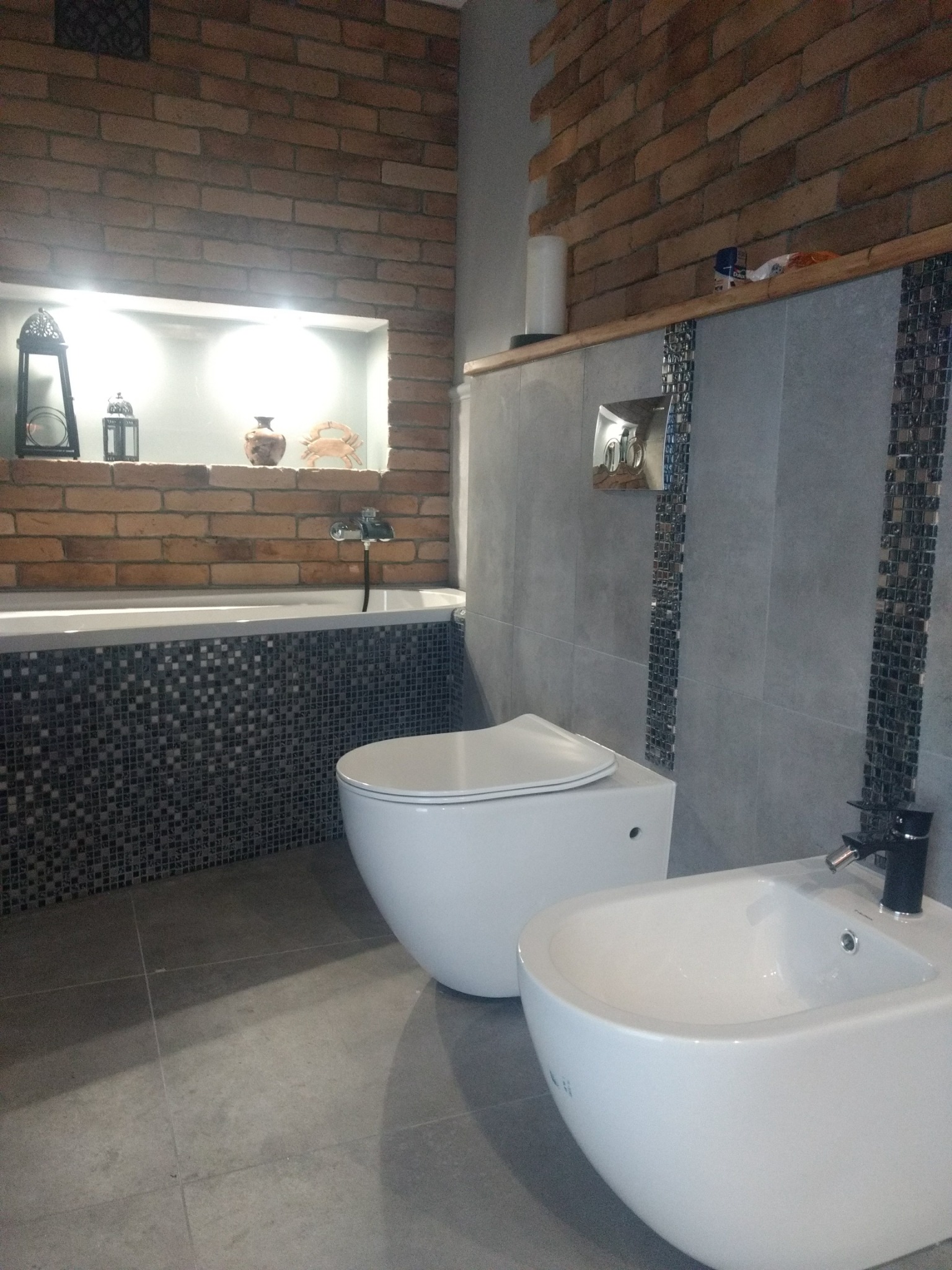 Nowoczesna łazienka z białym bidetem i toaletą, wykończona cegłą i szarą płytką z mozaikowymi akcentami.