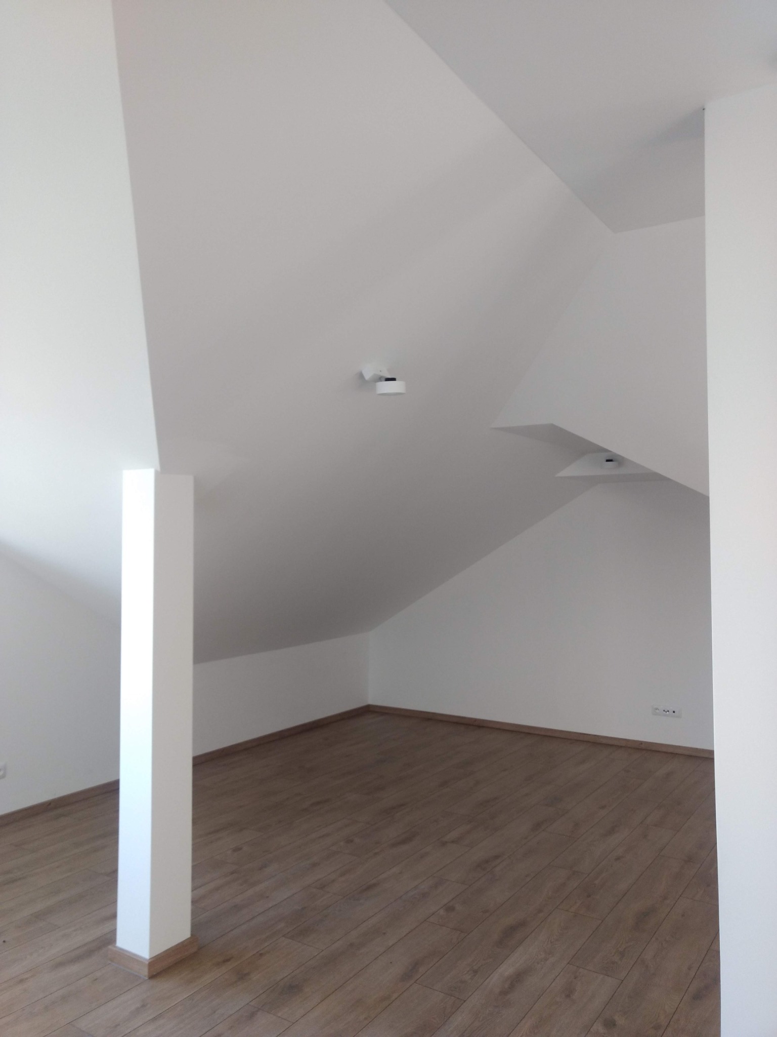 Poddasze po remoncie z drewnianą podłogą, białe ściany i kolumna nośna, widoczne lampy sufitowe.