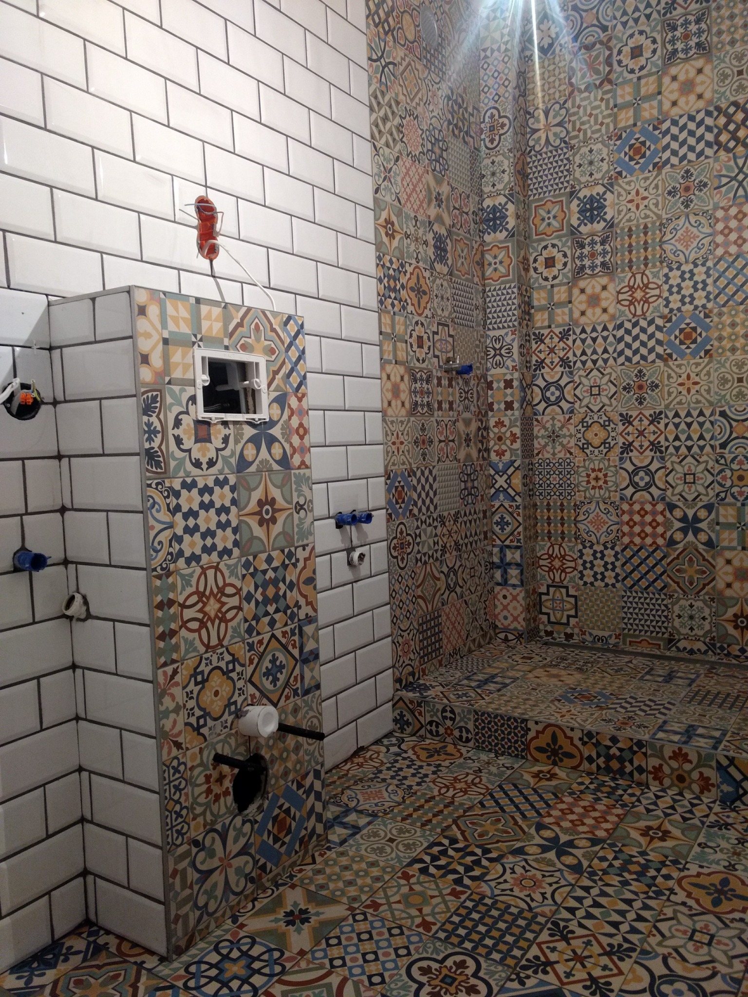 Wnętrze łazienki w trakcie wykańczania z białą cegiełkową glazurą i dekoracyjną mozaiką na ścianach i podłodze, widoczne wystające instalacje hydrauliczne i elektryczne.