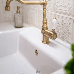 Biała, ceramiczna umywalka z mosiężną armaturą w stylu retro, dekoracyjne płytki ścienne z motywem kwiatowym, dozownik do mydła w tle, logo firmy w prawym dolnym rogu.