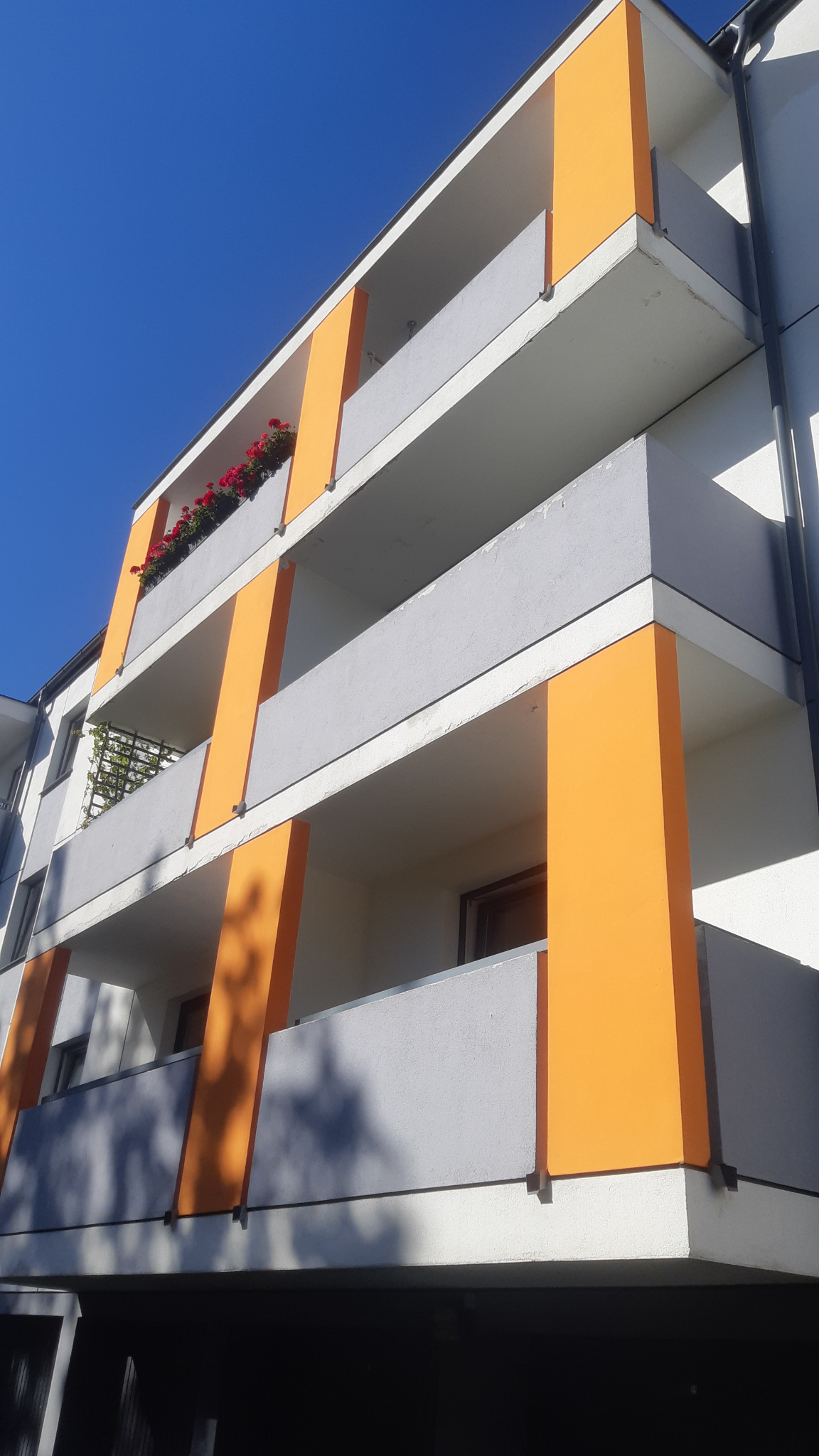 Wielopiętrowy budynek z balkonami, pomalowanymi na szaro z pomarańczowymi akcentami. Na balkonie czerwone kwiaty w donicach. Ujęcie z dołu, błękitne niebo w tle.