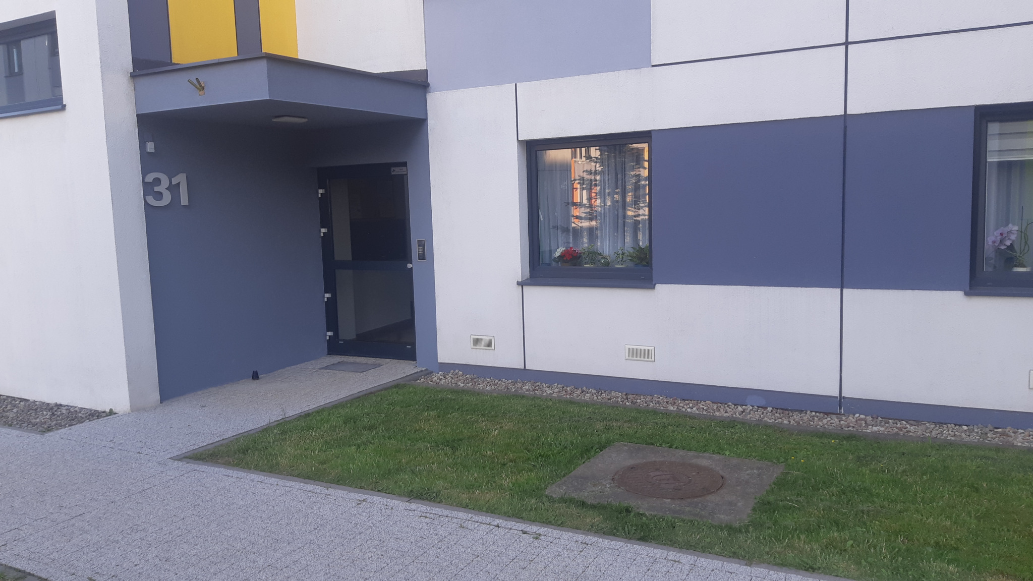Wejście do budynku z numerem 31, elewacja w odcieniach bieli i szarości, okna z kwiatami, trawnik. Widok na odnowioną fasadę budynku.