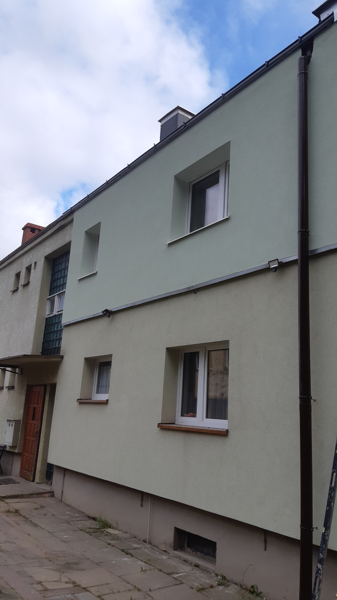 Odnowiona elewacja dwukondygnacyjnego budynku z nową, dwukolorową fasadą w odcieniach zieleni i beżu, z widoczną rynną i fragmentem drabiny.