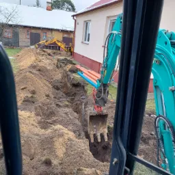 Wykopany rów z ułożonymi rurami, widok z kabiny turkusowej koparki, w tle żółta koparka JCB przy budynku.