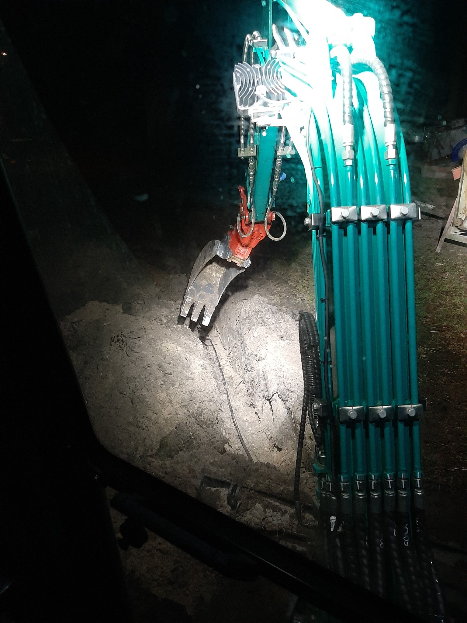 Nocny widok z kabiny koparki na oświetlony wykop, z widoczną łyżką i hydraulicznymi przewodami w kolorze turkusowym.