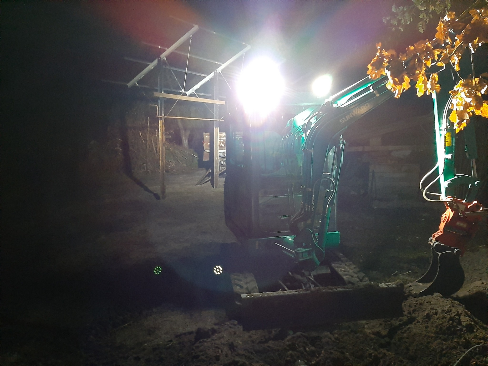 Nocny widok na małą, zieloną koparkę SUNWARD w trakcie pracy przy oświetlonym wykopie, z widoczną konstrukcją w tle i gałęzią drzewa z żółtymi liśćmi po prawej stronie.