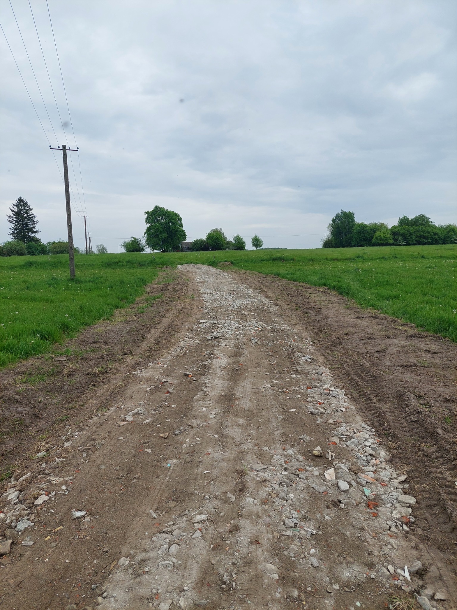 Utrwalona droga gruntowa z gruzem i śladami opon w wiejskim krajobrazie, słupy elektryczne w oddali pod pochmurnym niebem.