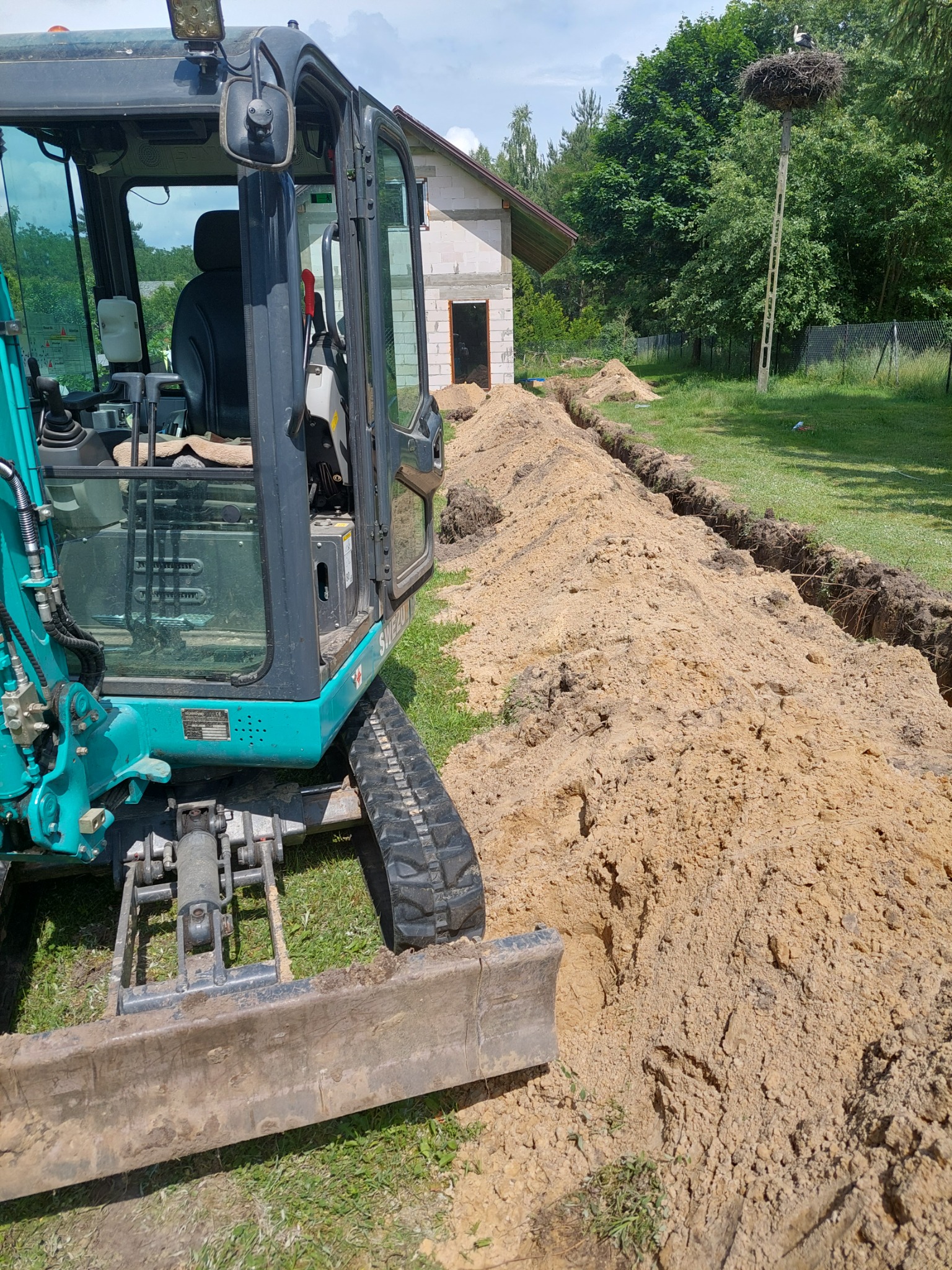 Turkusowa mini-koparka obok wykopanego, długiego rowu w ziemi na trawiastym terenie; w tle budynek w stanie surowym i gniazdo bocianie na słupie.