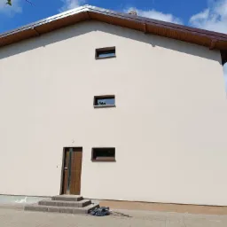 Elewacja dwukondygnacyjnego domu po renowacji, z brązowymi oknami i drzwiami wejściowymi. Na chodniku leżą narzędzia, a przy ścianie widać rusztowanie.