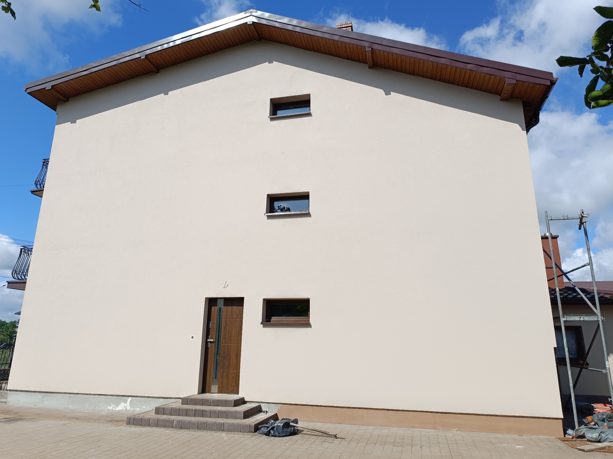 Elewacja dwukondygnacyjnego domu po renowacji, z brązowymi oknami i drzwiami wejściowymi. Na chodniku leżą narzędzia, a przy ścianie widać rusztowanie.