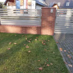 Nowoczesna brama wjazdowa z poziomych profili metalowych, osadzona na murowanych słupkach z czerwonej cegły, z widocznym domofonem na jednym ze słupków, otoczona trawnikiem z opadłymi liśćmi.