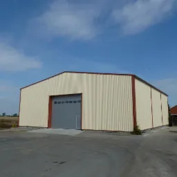 Gotowa hala stalowa z kremowymi ścianami obłożonymi blachą trapezową i szarą bramą segmentową, widoczna na tle pola i błękitnego nieba z pojedynczymi obłokami.