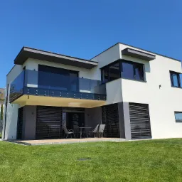 Nowoczesny dom z białymi ścianami, szarymi akcentami i szklaną balustradą na balkonie, widok z zadbanego trawnika w słoneczny dzień.