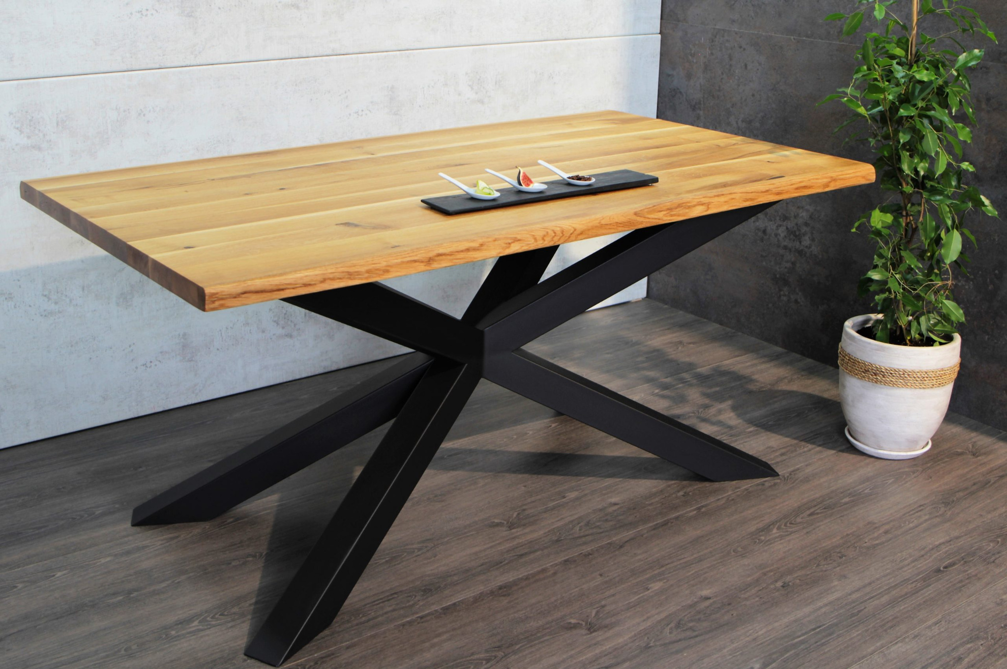 Stół z blatem z jasnego drewna z nieregularną krawędzią i czarną, geometryczną podstawą w kształcie gwiazdy, na blacie czarna deska z trzema przekąskami na białych łyżeczkach, w tle roślina...