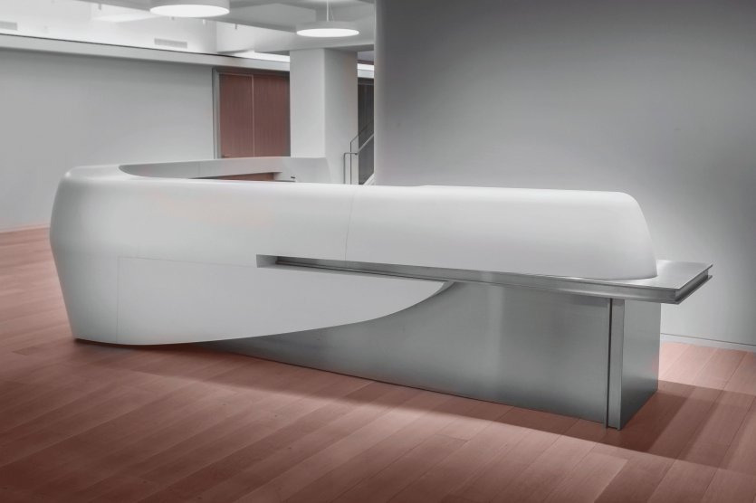 Nowoczesna biała lada recepcyjna o zaokrąglonym kształcie z elementami ze stali nierdzewnej, umieszczona w minimalistycznym wnętrzu z drewnianą podłogą i białymi ścianami.