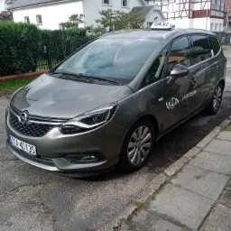 Szary minivan Opel Zafira z napisem 'TAXI' na dachu, zaparkowany na chodniku w Wisełce, z widocznym numerem telefonu na boku.