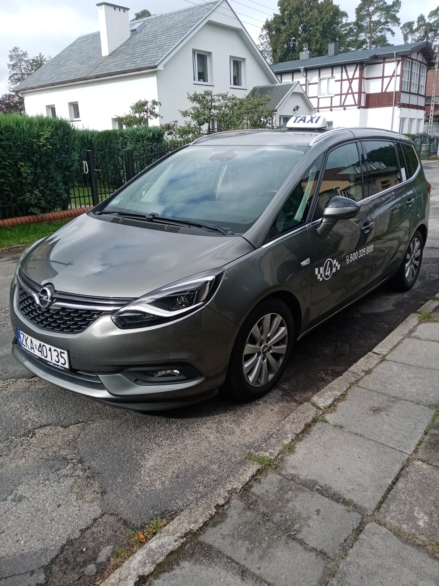 Szary minivan Opel Zafira z napisem 'TAXI' na dachu, zaparkowany na chodniku w Wisełce, z widocznym numerem telefonu na boku.