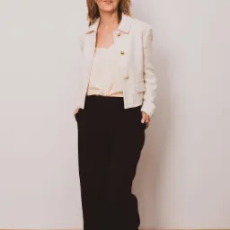 Kobieta w okularach prezentuje stylizację: kremowa bluzka, biała tweedowa marynarka z złotymi guzikami i czarne spodnie z szerokimi nogawkami, ręce w kieszeniach.