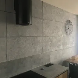 Szara ściana w kuchni z efektem betonu architektonicznego, podzielona na prostokątne panele, z czarnym okapem nad płytą i dekoracyjnym zegarem na ścianie.