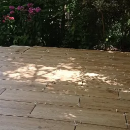 Układanie drewnopodobnych płytek na tarasie z widocznymi krzyżykami dystansowymi, w tle bujna roślinność ogrodowa i padające cienie drzew.